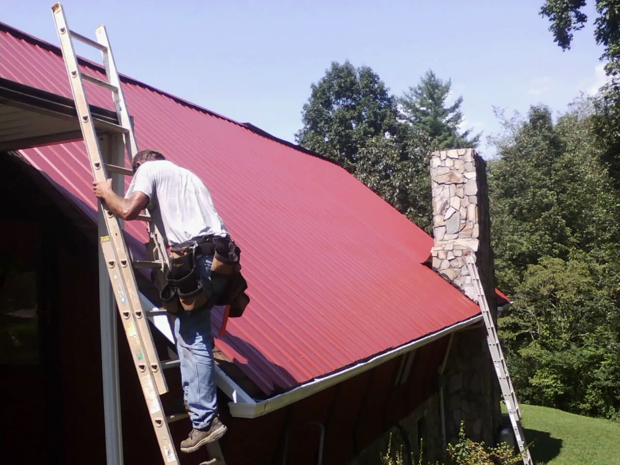 Roofer on a ladder beside a red metal roof and stone chimney — Soffit & Fascia Repair by Roofing Brothers in Paradise